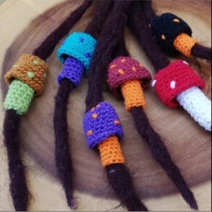 Op de afbeelding: Een verzameling kleurrijke gehaakte paddenstoel kralen, elk met een andere kleur voor de dop en de steel. De kralen zijn geregen aan bruine koorden, die lijken op dreadlocks.