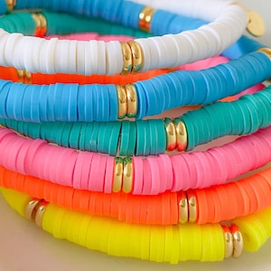 May include: A stack of colourful bracelets made of round, flat beads. The bracelets are in shades of white, blue, turquoise, pink, orange, and yellow. Each bracelet has a gold accent.