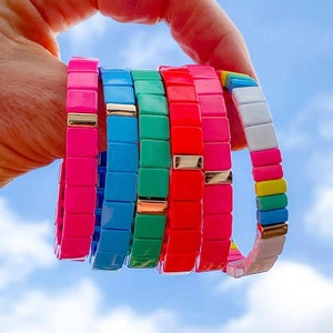 May include: A stack of colourful bracelets made of square tiles. The bracelets are in shades of pink, blue, green, and red. The bracelets have gold accents.
