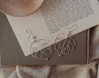 May include: A pair of silver monstera leaf earrings on a brown fabric surface. The earrings are delicate and have a simple design.