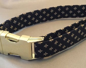 Puede incluir: Un collar de perro negro con una hebilla dorada y un patrón de flores de lis doradas.