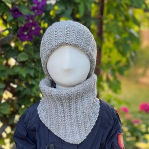 Könnte beinhalten: Eine hellgraue Strickmütze und ein passender gerippter Schlauchschal an einer Schaufensterpuppe. Das Set zeichnet sich durch ein durchgehendes, strukturiertes Strickmuster aus neutralem, meliertem grauen Garn aus, das Kopf und Hals bedeckt.