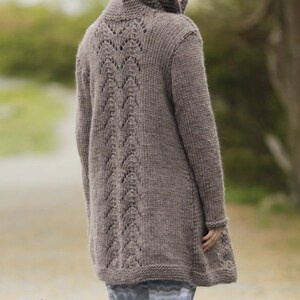 May include: A brown hooded cardigan with a lacy back detail. The cardigan is long and has a relaxed fit. The model is wearing the cardigan with a pair of striped pants. DROPS DESIGN www.garnstudio.com
