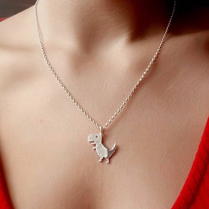 May include: A silver necklace with a small, pixelated dinosaur pendant. The necklace is worn by a person wearing a red shirt.