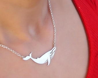 Op de afbeelding: Een zilveren ketting met een walvis hanger. De walvis kijkt naar rechts en heeft een gedetailleerd ontwerp. De ketting wordt gedragen door iemand die een rood shirt draagt.