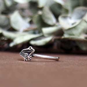 Puede incluir: Un anillo de nariz de plata con forma de rana con un diseño simple. El anillo está sobre una superficie marrón.