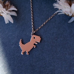 May include: A silver chain necklace with a pixelated dinosaur pendant. The pendant is a silver color and is shaped like the classic video game dinosaur.