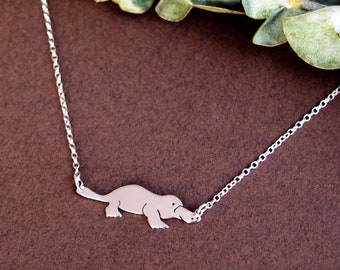 Peut inclure: Un collier en chaîne argent avec un pendentif en forme d'ornithorynque argenté. Le collier est sur un fond brun.