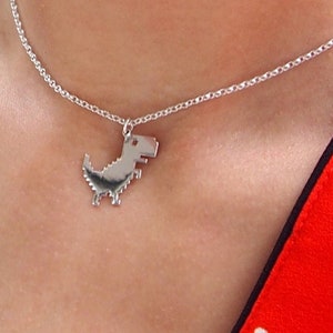 May include: A silver necklace with a pixelated dinosaur pendant. The necklace is worn by a person wearing a red and white polka dot shirt.