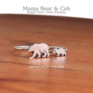 Peut inclure: Bague en argent représentant une maman ours et son ourson. Le texte "Mama Bear & Cub Build Your Own Family" et "Add one cub for each child" est visible. La bague est posée sur une surface en bois.