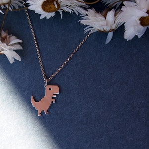 May include: A silver chain necklace with a pixelated dinosaur pendant. The necklace is laying on a blue surface with white flowers in the background.