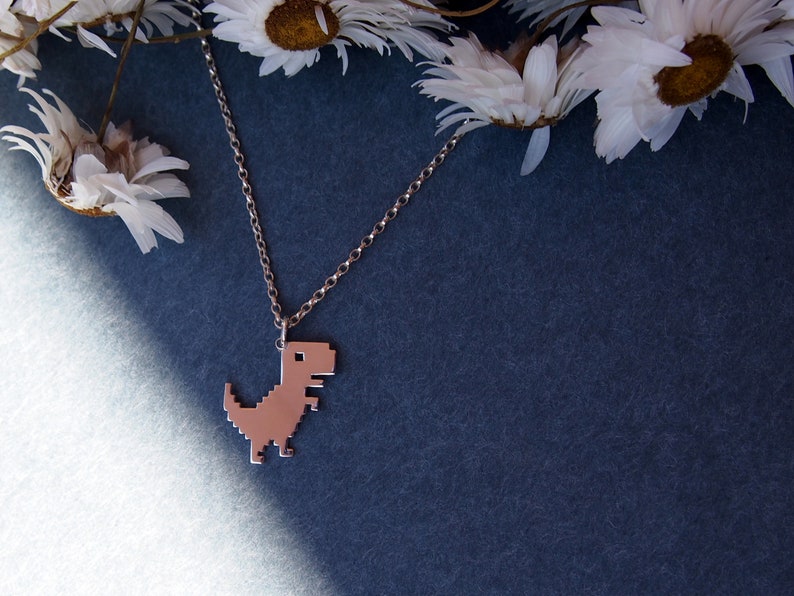May include: A silver chain necklace with a pixelated dinosaur pendant. The necklace is laying on a blue surface with white flowers in the background.
