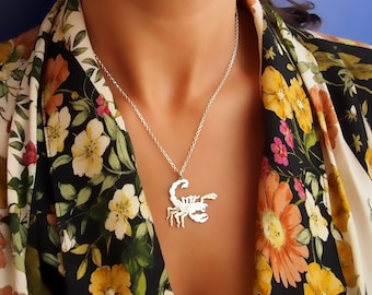以下が含まれることがあります： サソリのペンダントが付いたシルバーネックレス。ネックレスは、花柄のシャツを着た人が着用しています。
