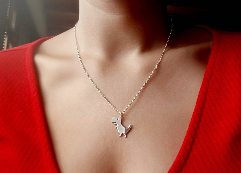 May include: A silver necklace with a small, pixelated dinosaur pendant. The necklace is worn by a person wearing a red shirt.
