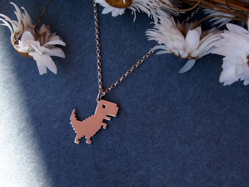 May include: A silver chain necklace with a pixelated dinosaur pendant. The pendant is a silver color and is shaped like the classic video game dinosaur.
