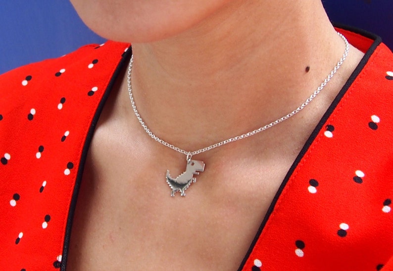 May include: A silver necklace with a pixelated dinosaur pendant. The necklace is worn by a person wearing a red and white polka dot shirt.