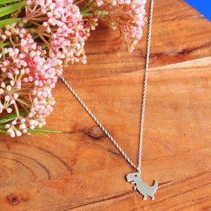 May include: A silver chain necklace with a pixelated dinosaur pendant. The necklace is on a wooden surface with pink flowers.
