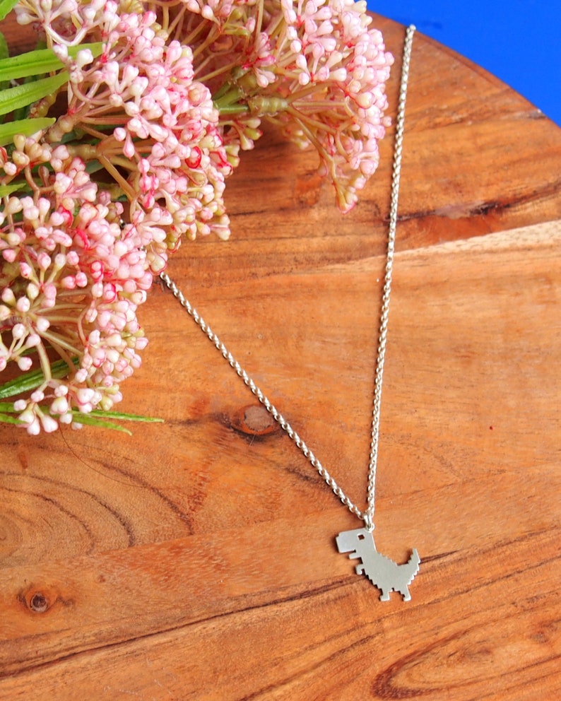 May include: A silver chain necklace with a pixelated dinosaur pendant. The necklace is on a wooden surface with pink flowers.