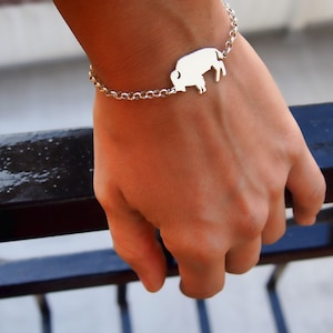 Op de afbeelding: Een zilveren kettingarmband met een buffel-silhouetbedel. De armband zit om een pols.