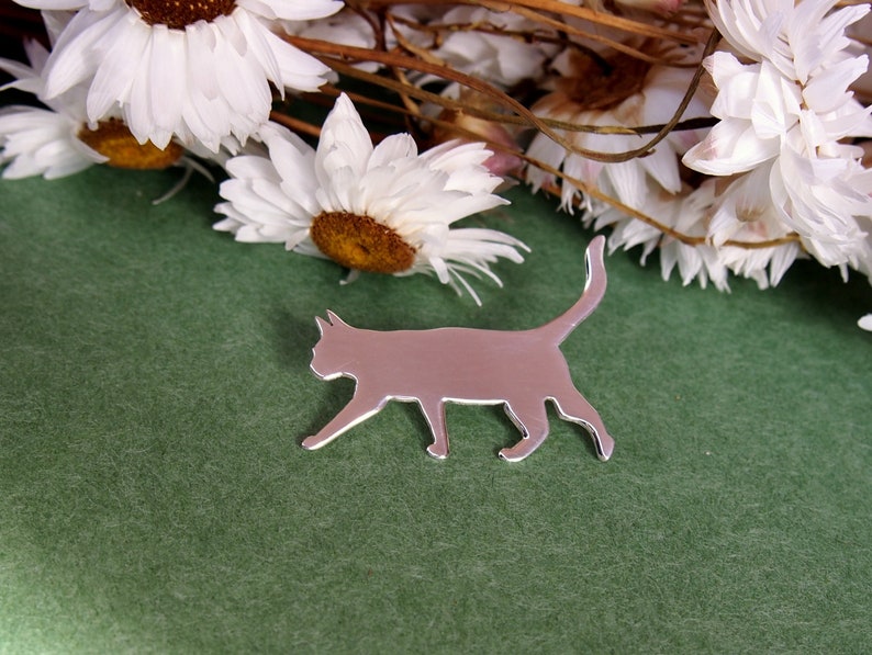 K&ouml;nnte beinhalten: Ein silberfarbener Katzen-Silhouetten-Anh&auml;nger auf gr&uuml;nem Hintergrund mit getrockneten wei&szlig;en Blumen im Hintergrund.