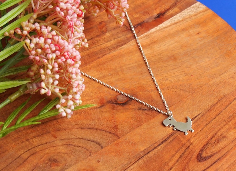 May include: A silver necklace with a pixelated dinosaur pendant. The necklace is on a wooden surface with a pink flower.