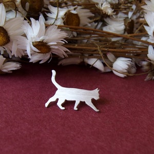 K&ouml;nnte beinhalten: Eine silberne Brosche mit einer Katzen-Silhouette auf einem burgunderroten Hintergrund mit getrockneten wei&szlig;en G&auml;nsebl&uuml;mchen.