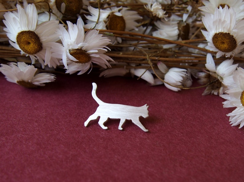 K&ouml;nnte beinhalten: Eine silberne Brosche mit einer Katzen-Silhouette auf einem burgunderroten Hintergrund mit getrockneten wei&szlig;en G&auml;nsebl&uuml;mchen.