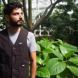 K&ouml;nnte beinhalten: Ein Mann tr&auml;gt eine braune Weste mit einer kleinen Katzennadel &uuml;ber einem grauen T-Shirt. Er steht in einem &uuml;ppigen gr&uuml;nen Garten mit gro&szlig;en Bl&auml;ttern.