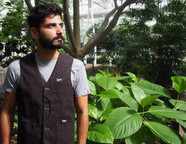 K&ouml;nnte beinhalten: Ein Mann tr&auml;gt eine braune Weste mit einer kleinen Katzennadel &uuml;ber einem grauen T-Shirt. Er steht in einem &uuml;ppigen gr&uuml;nen Garten mit gro&szlig;en Bl&auml;ttern.