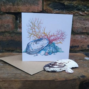 Puede incluir: Una tarjeta de felicitación con una ilustración de acuarela de una concha, coral y algas marinas. La concha es blanca con acentos azules y verdes. El coral es rojo y naranja. Las algas marinas son amarillas y verdes.