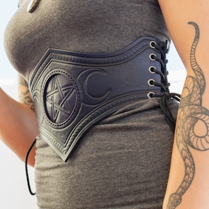 May include: A grey shirt with a black leather belt featuring a pentagram and crescent moon design. The belt has a lace-up back and is worn over a grey shirt. The person wearing the belt has a snake tattoo on their arm.