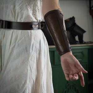 Puede incluir: Un protector de brazo de cuero marrón con un patrón texturizado se lleva en un brazo. El protector de brazo está asegurado con remaches y tiene una forma curva. También se ve un cinturón de cuero marrón con una hebilla dorada.
