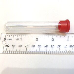 May include: A clear, cylindrical test tube with a red cap, approximately 4 inches long, is positioned next to a ruler. The ruler shows inch measurements. The tube is empty and made of transparent material.