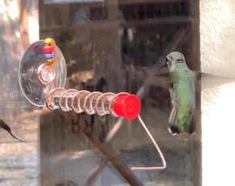 May include: A window-mounted hummingbird feeder with a clear suction cup, copper coil, and red feeding port. A small, green hummingbird is perched nearby. The feeder is designed to attract hummingbirds.