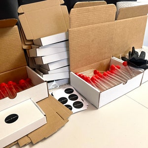 May include: An assortment of white boxes, cardboard boxes, and test tubes with red caps. The boxes are open, revealing the contents. Black gloves and stickers are also visible. The boxes are arranged on a white surface.