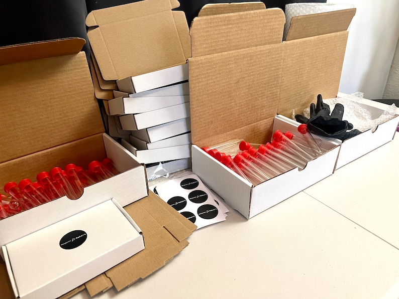 May include: An assortment of white boxes, cardboard boxes, and test tubes with red caps. The boxes are open, revealing the contents. Black gloves and stickers are also visible. The boxes are arranged on a white surface.