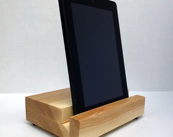 May include: A black tablet computer is positioned vertically in a light-colored wooden stand. The stand features a rectangular base with a grooved slot designed to hold the device securely in an upright position, showcasing natural wood grain.