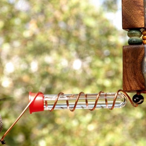 May include: A decorative hummingbird feeder with a clear glass tube and a red feeding port. The feeder is suspended from a wooden and glass structure with decorative beads. A hummingbird approaches the feeder.