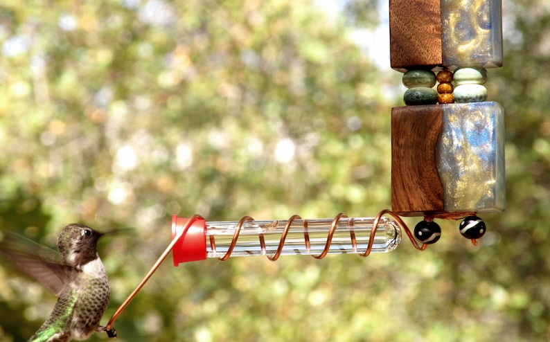 May include: A decorative hummingbird feeder with a clear glass tube and a red feeding port. The feeder is suspended from a wooden and glass structure with decorative beads. A hummingbird approaches the feeder.