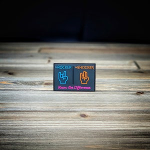 K&ouml;nnte beinhalten: Ein schwarzer Patch mit den Worten "The Rocker" und "The Shocker" in wei&szlig;er Schrift. Der Patch zeigt zwei Handgesten, eine in Blau und eine in Orange, mit dem Text "Know the Difference" in Pink darunter.