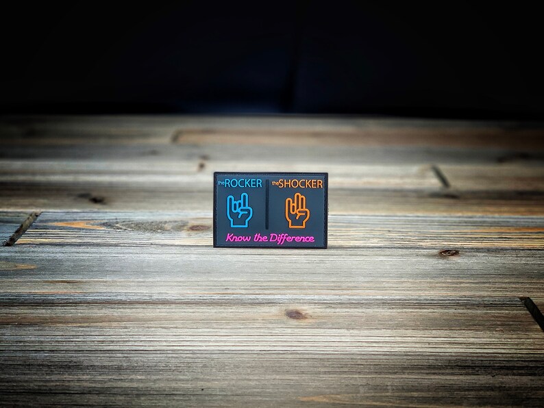 K&ouml;nnte beinhalten: Ein schwarzer Patch mit den Worten "The Rocker" und "The Shocker" in wei&szlig;er Schrift. Der Patch zeigt zwei Handgesten, eine in Blau und eine in Orange, mit dem Text "Know the Difference" in Pink darunter.