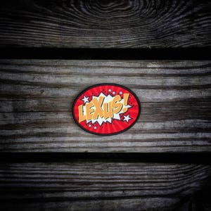 May include: A red and yellow comic book style patch with the word "LEXUS!" in a white speech bubble. The patch is on a wooden background.