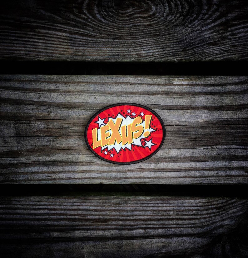 May include: A red and yellow comic book style patch with the word "LEXUS!" in a white speech bubble. The patch is on a wooden background.