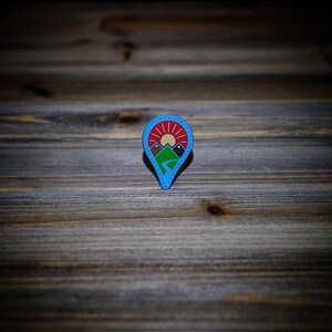 May include: A blue enamel pin with a white outline depicting a mountain range with a red sun rising behind it. The pin is shaped like a location marker.