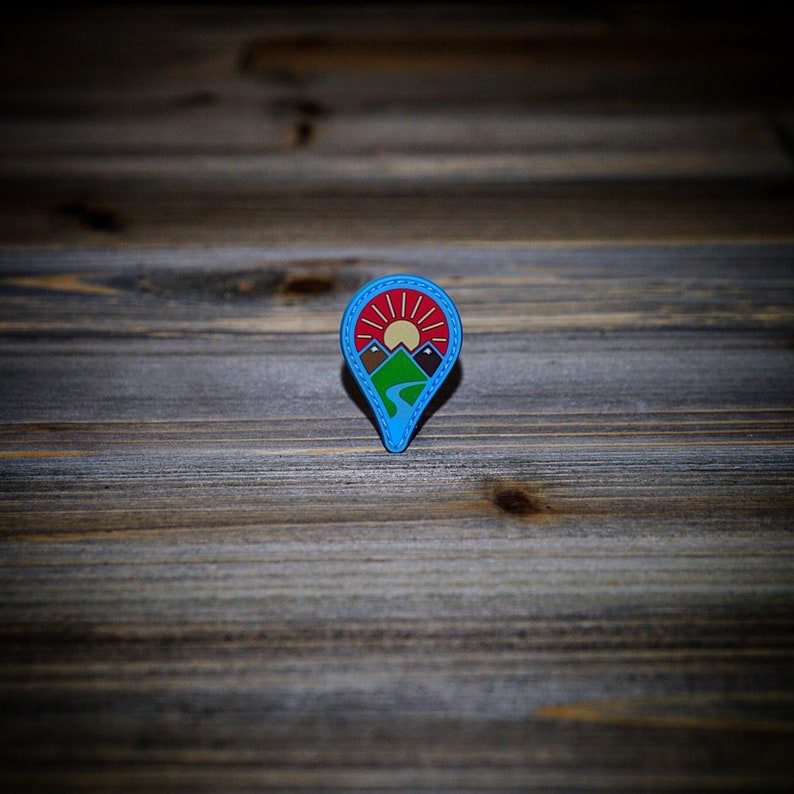 May include: A blue enamel pin with a white outline depicting a mountain range with a red sun rising behind it. The pin is shaped like a location marker.