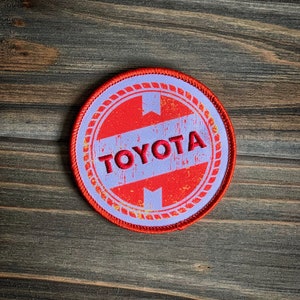 May include: A red and white circular patch with a white border and a rope design. The patch has the word "TOYOTA" in red lettering.