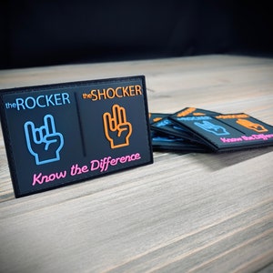 K&ouml;nnte beinhalten: Schwarzer Patch mit dem Text "the ROCKER" und "the SHOCKER" mit einem blauen und orangefarbenen Handgesten-Symbol neben jedem. Der Text "Know the Difference" befindet sich unter den Symbolen in Pink.