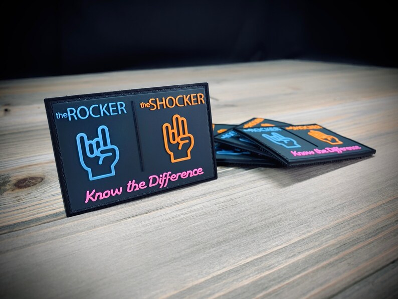 K&ouml;nnte beinhalten: Schwarzer Patch mit dem Text "the ROCKER" und "the SHOCKER" mit einem blauen und orangefarbenen Handgesten-Symbol neben jedem. Der Text "Know the Difference" befindet sich unter den Symbolen in Pink.