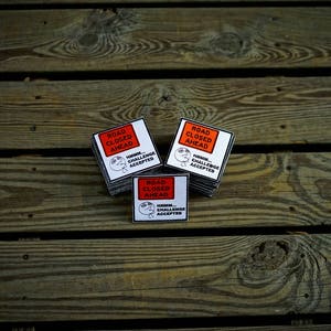 Op de afbeelding: Drie witte vierkante stickers met zwarte tekst en een cartoon gezicht. De stickers zeggen "Weg afgesloten" en "Hmm... Uitdaging geaccepteerd".