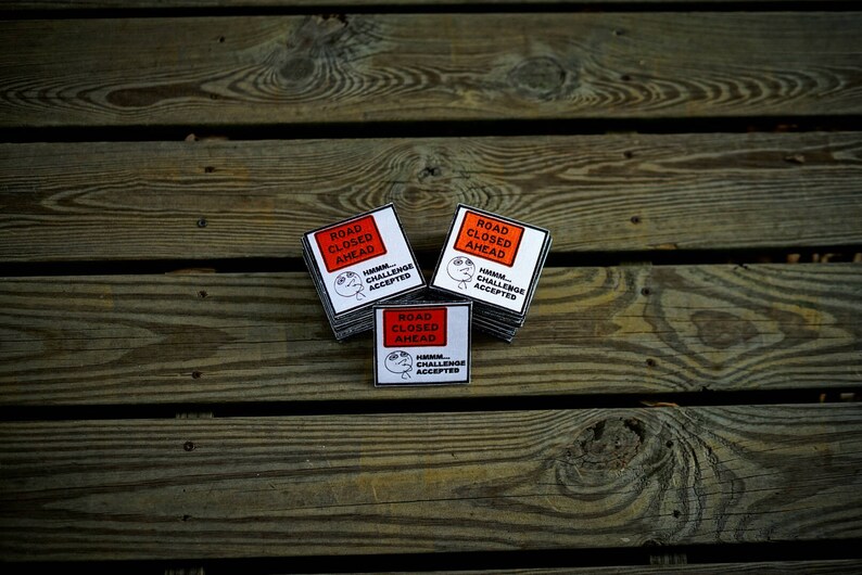 Op de afbeelding: Drie witte vierkante stickers met zwarte tekst en een cartoon gezicht. De stickers zeggen "Weg afgesloten" en "Hmm... Uitdaging geaccepteerd".